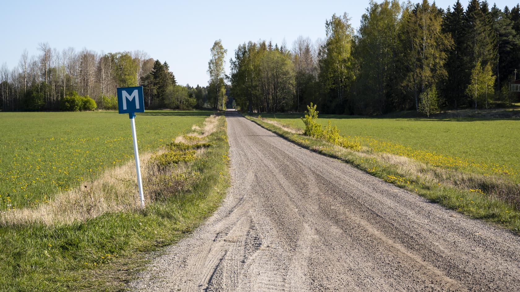 En grusväg mellan två åkrar. I vägkanten står en blå skylt med bokstaven M för mötesplats.