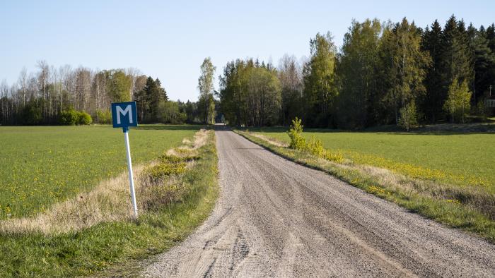 En grusväg mellan två åkrar. I vägkanten står en blå skylt med bokstaven M för mötesplats.