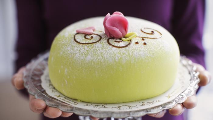 Någon håller i ett tårtfat med en grön prinsesstårta med rosa marsipanros på toppen.