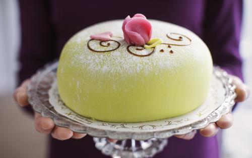 Någon håller i ett tårtfat med en grön prinsesstårta med rosa marsipanros på toppen.