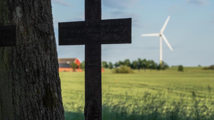 Ett metallkors står på en gravsten i skuggan av ett gammalt träd. I bakgrunden syns ett vindkraftverk.