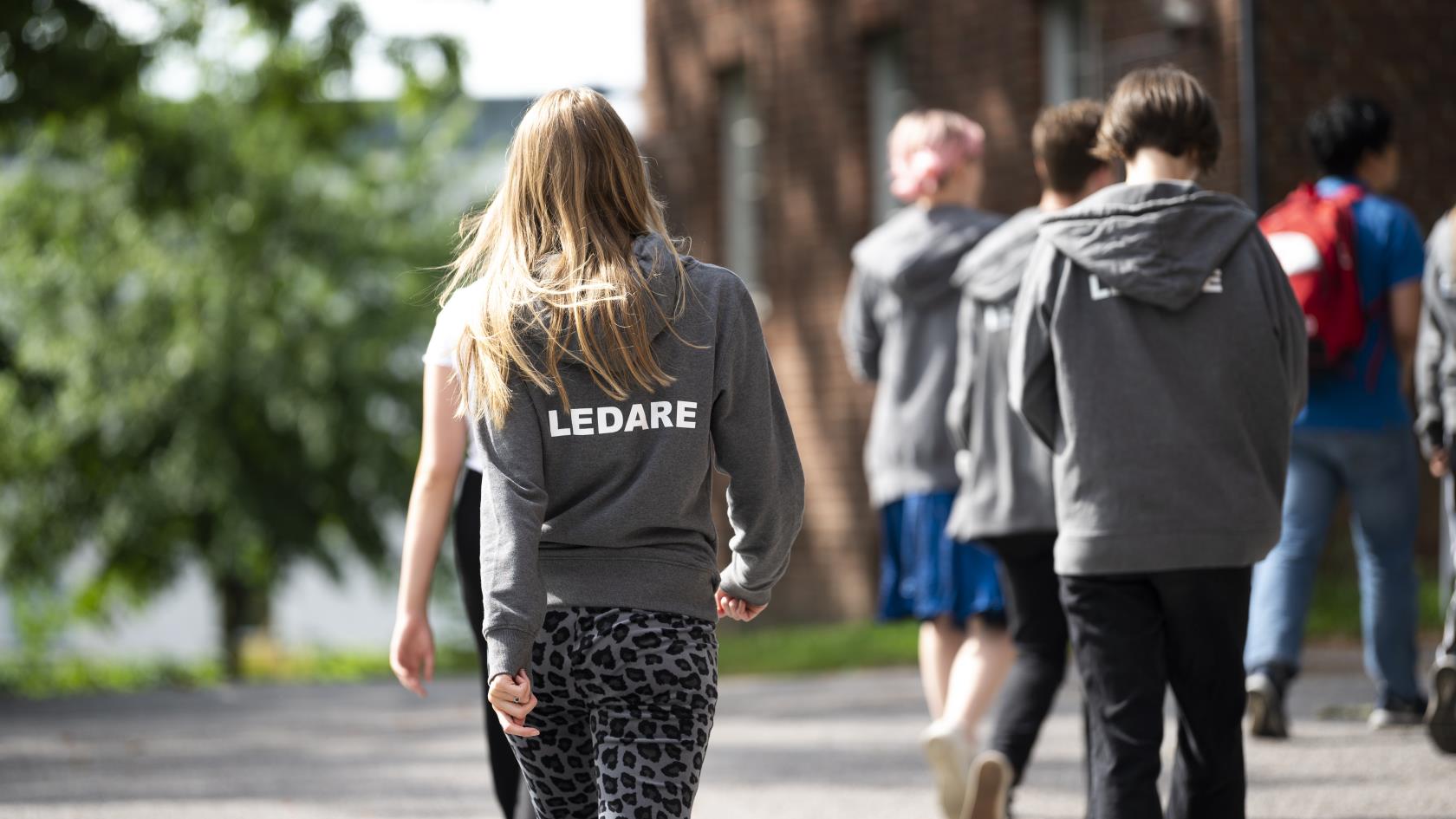 Vi ser ryggtavlorna på ett antal ungdomar. På en av deras huvtröjor står texten "Ledare".