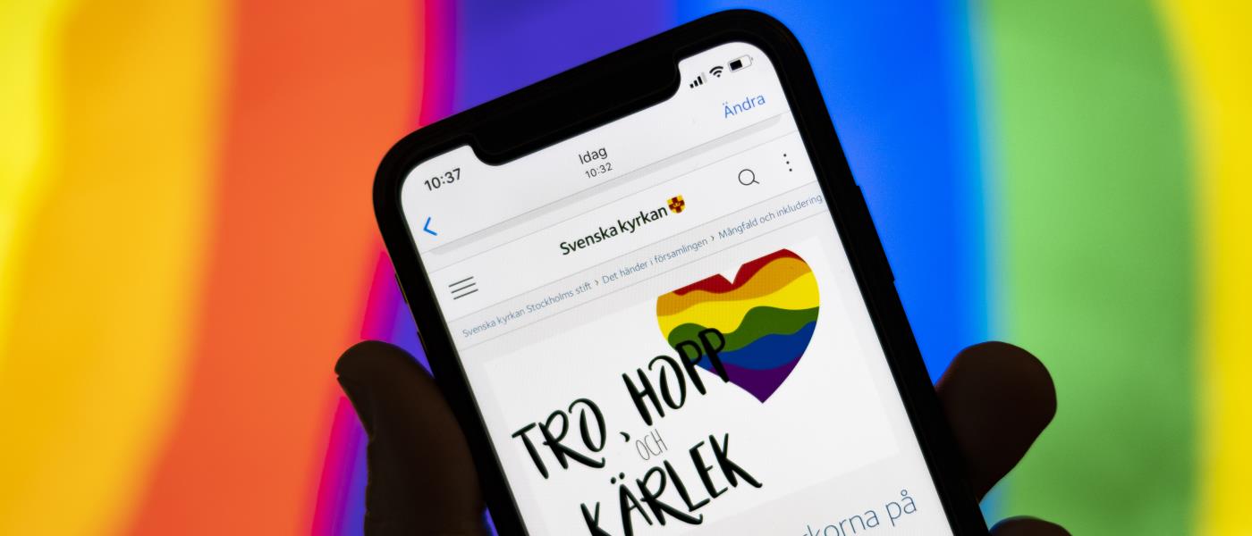 Någon håller en mobiltelefon där skärmen visar Svenska kyrkans information om Pride. I bakgrunden syns regnbågsfärgerna.