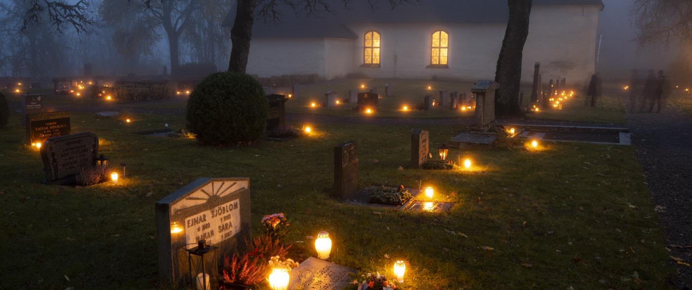 Höstkväll på kyrkogård där varje grav har varsitt tänt ljus. Det lyser inne i kyrkan.