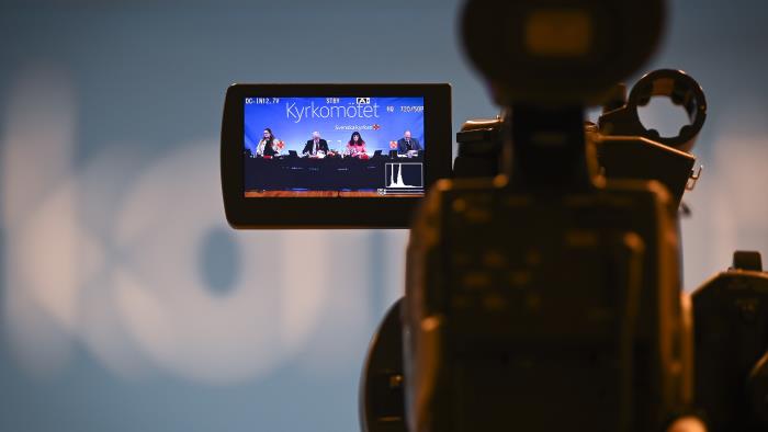 På skärmen till en videokamera syns en panel med fyra personer framför en fond med texten Kyrkomötet.