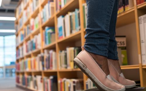 Någon i rosa tygskor står på tå på en pall inne på ett bibliotek.