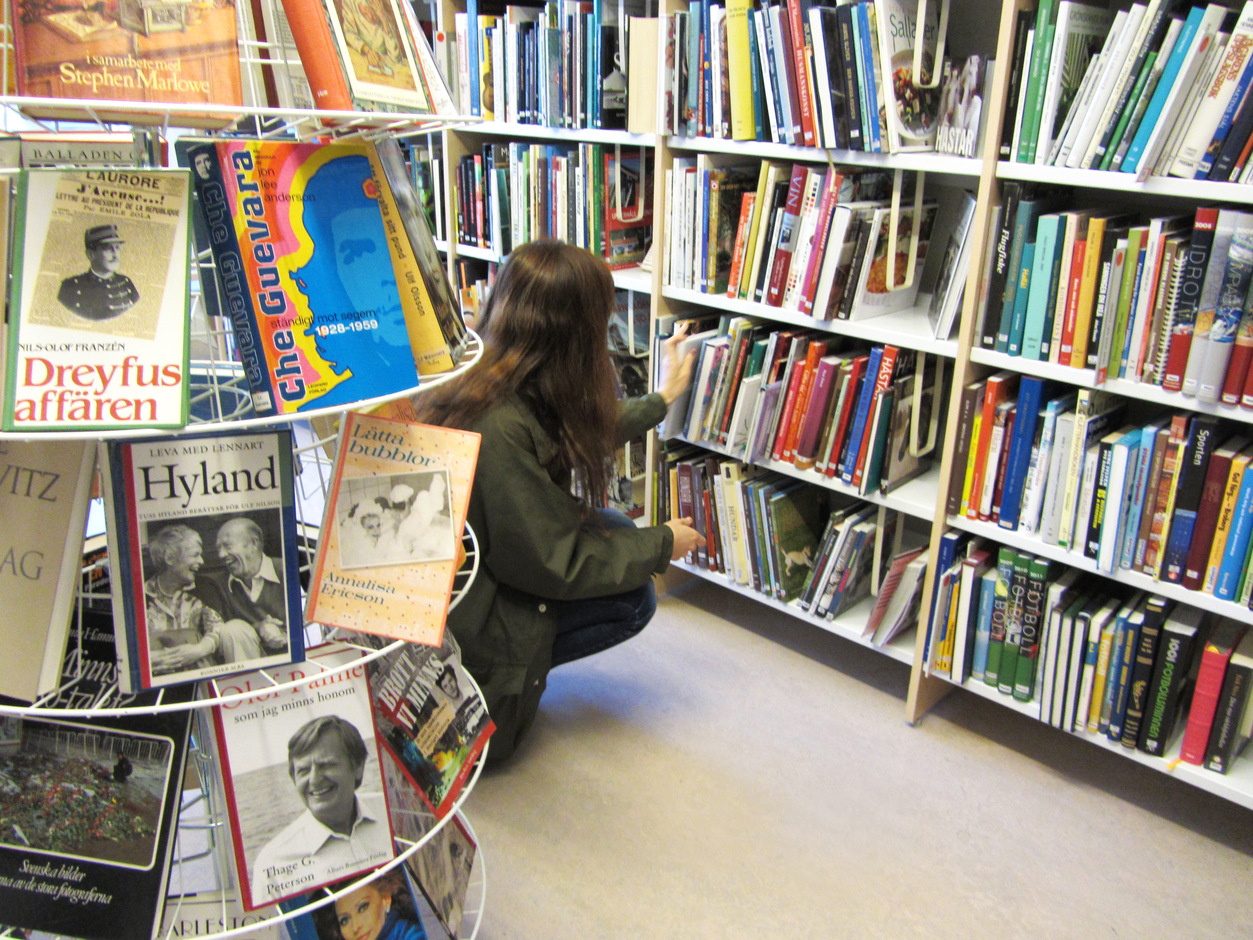 En kvinna sitter på huk och bläddrar bland böcker i en bokhylla på ett bibliotek.