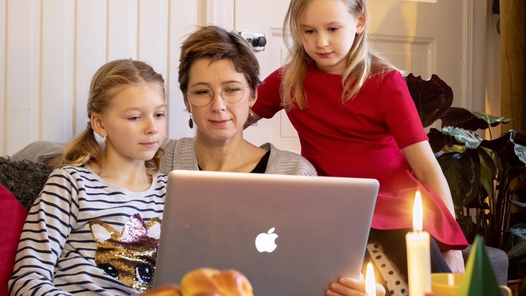 En kvinna och hennes två döttrar sitter med en bärbar dator i knät vid ett uppdukat adventsfika.