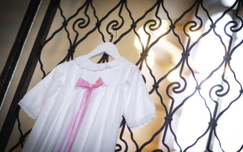 En vit dopklänning med rosa rosett hänger på en metallgrind i kyrkan.