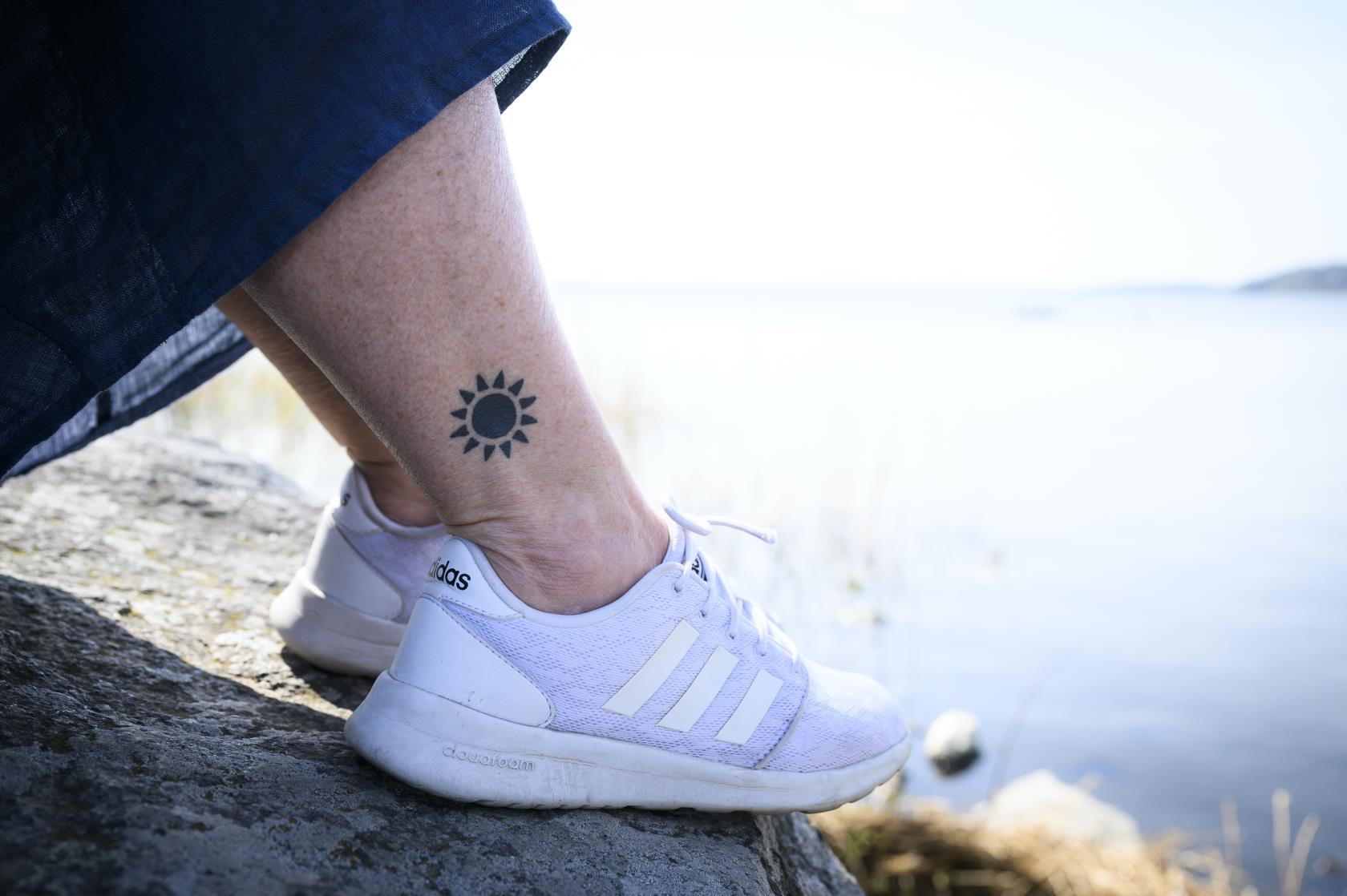 Någon sitter på en klippa vid vattnet en sommardag. Personen har en sol tatuerad på vaden.