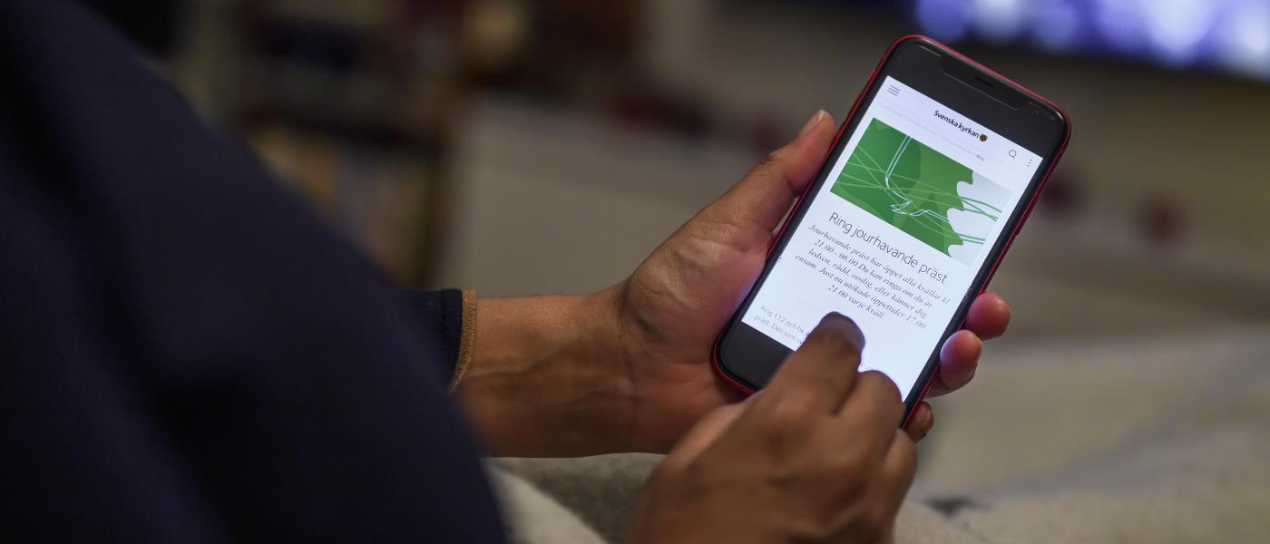 En person har sin mobiltelefon i händerna. Skärmen visar Svenska kyrkans information om jourhavande präster.