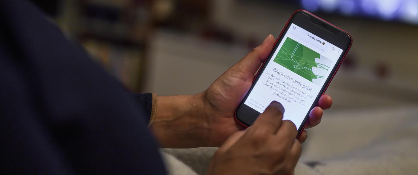 En person har sin mobiltelefon i händerna. Skärmen visar Svenska kyrkans information om jourhavande präster.