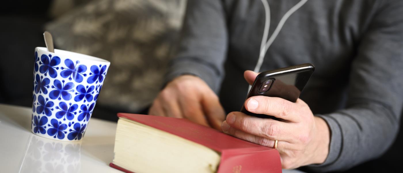 En präst sitter med hörlurar och pratar i mobiltelefon. En psalmbok ligger på bordet framför.