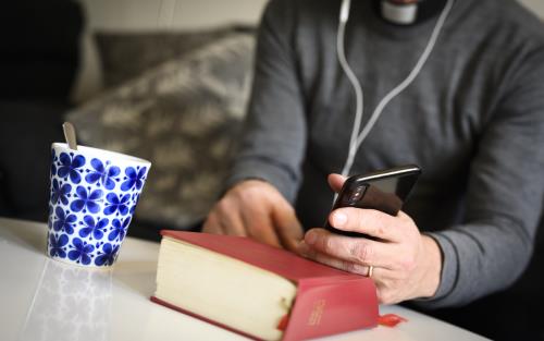 En präst sitter med hörlurar och pratar i mobiltelefon. En psalmbok ligger på bordet framför.