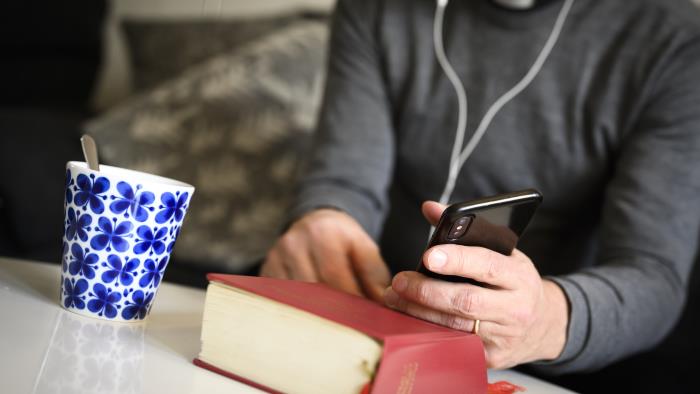 En präst sitter med hörlurar och pratar i mobiltelefon. En psalmbok ligger på bordet framför.