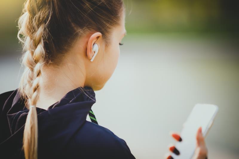 En tjej med fläta och hörlurar står utomhus med mobilen i handen.