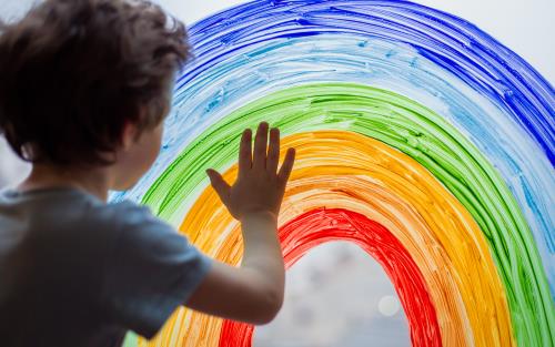 En liten pojke håller handen på en regnbåge målad på ett fönster.