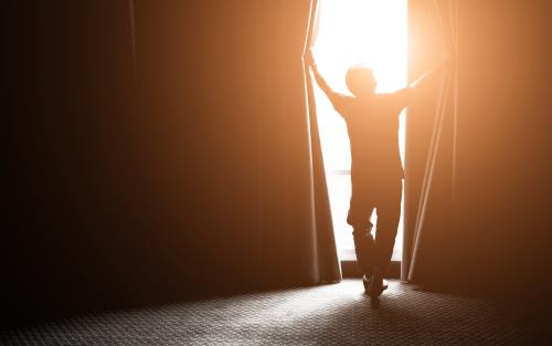Siluetten av en person som öppnar upp gardinerna vid ett stort fönster, solen lyser in.