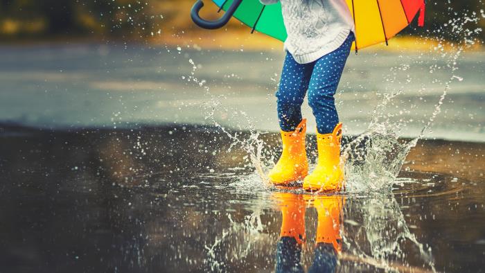 Ett barn med ett färgglatt paraply i handen och gula gummistövlar hoppar i en vattenpöl.