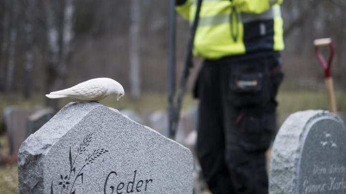 En keramik-fågel på en gravsten. I bakgrunden står en kyrkogårdsarbetare.