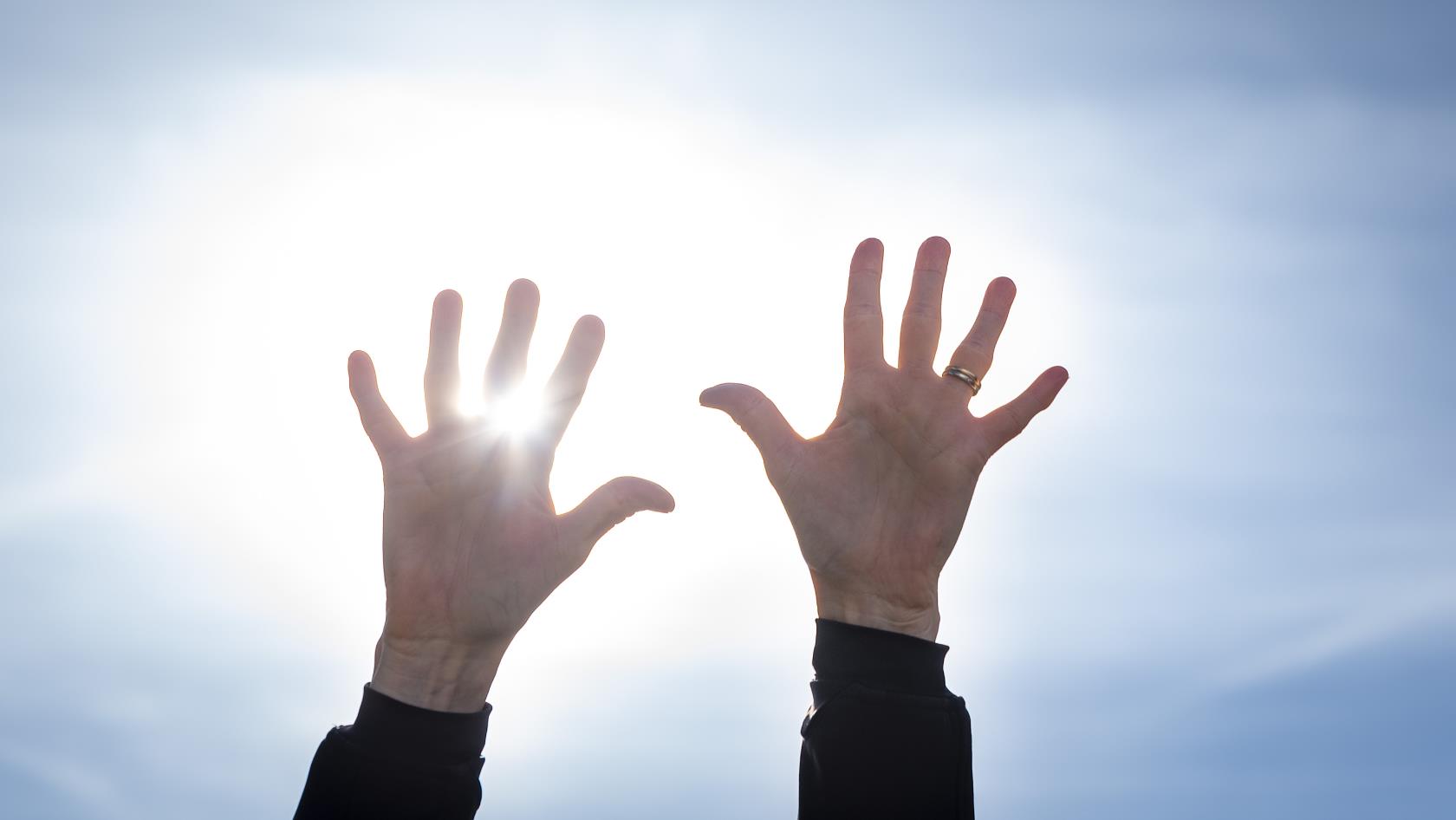 Två händer uppsträckta mot en blå himmel. Solen lysen mellan fingrarna.