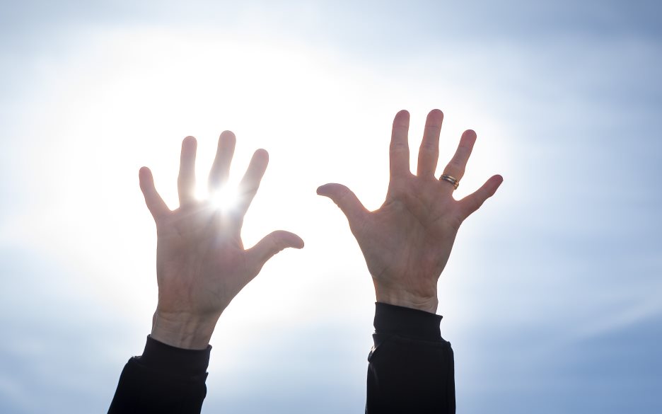 Två händer uppsträckta mot en blå himmel. Solen lysen mellan fingrarna.