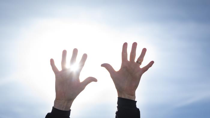 Två händer uppsträckta mot en blå himmel. Solen lysen mellan fingrarna.