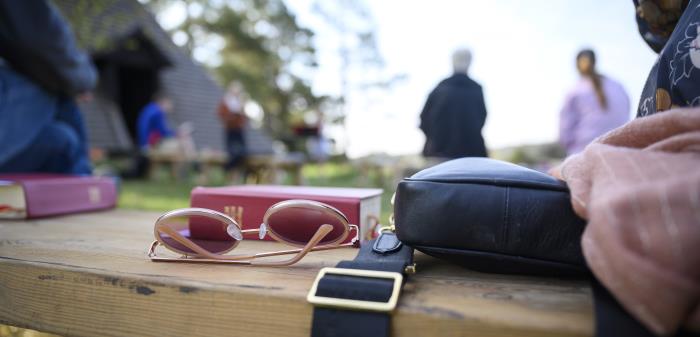 Ett par solglasögon och några psalmböcker ligger på en bänk utomhus. Människor syns i bakgrunden.