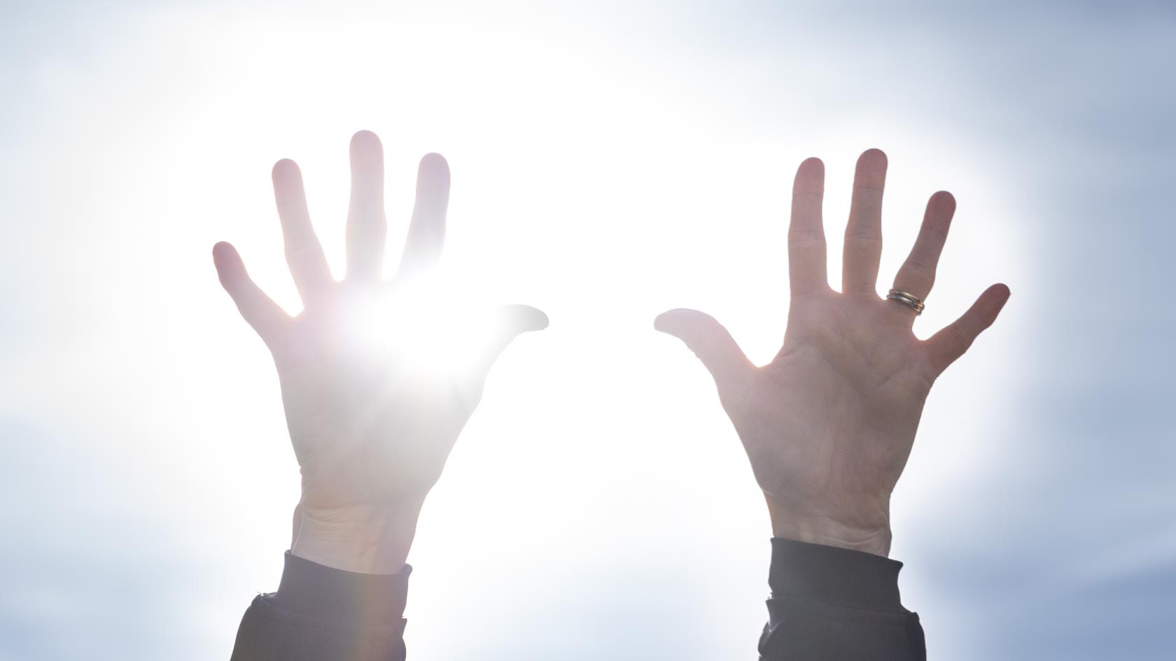 Två händer uppsträckta mot en blå himmel. Solen lysen mellan fingrarna.