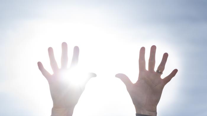 Två händer uppsträckta mot en blå himmel. Solen lysen mellan fingrarna.