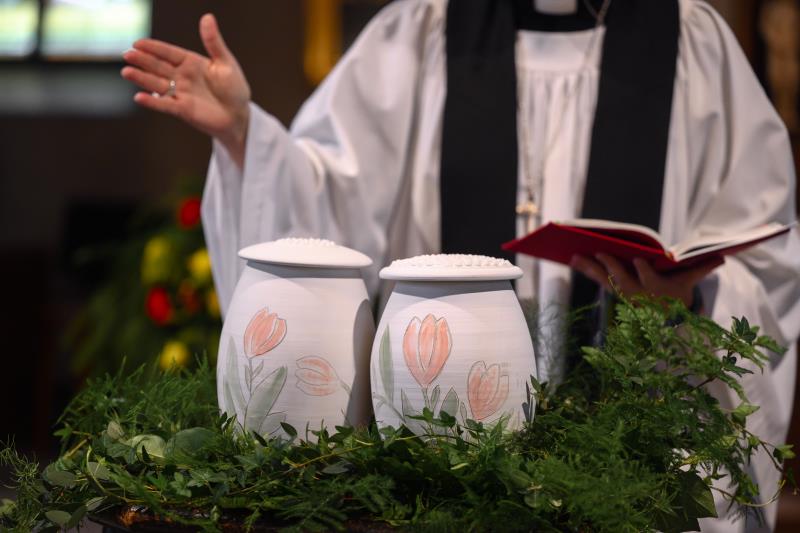 Två urnor med handmålade tulpaner står bland ormbunksblad på ett bord i kyrkan. En präst står bakom.