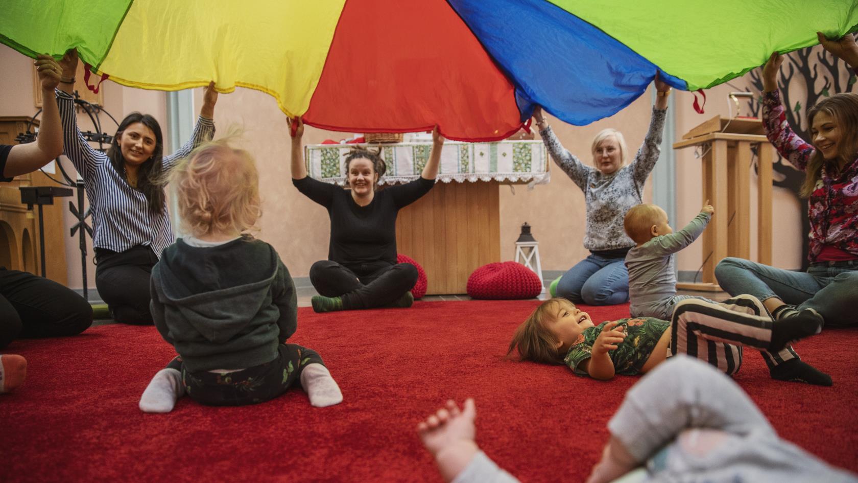 En grupp kvinnor sitter i en ring och lyfter tillsammans ett färgglatt tak. Inne i ringen, under taket, leker små bebisar.