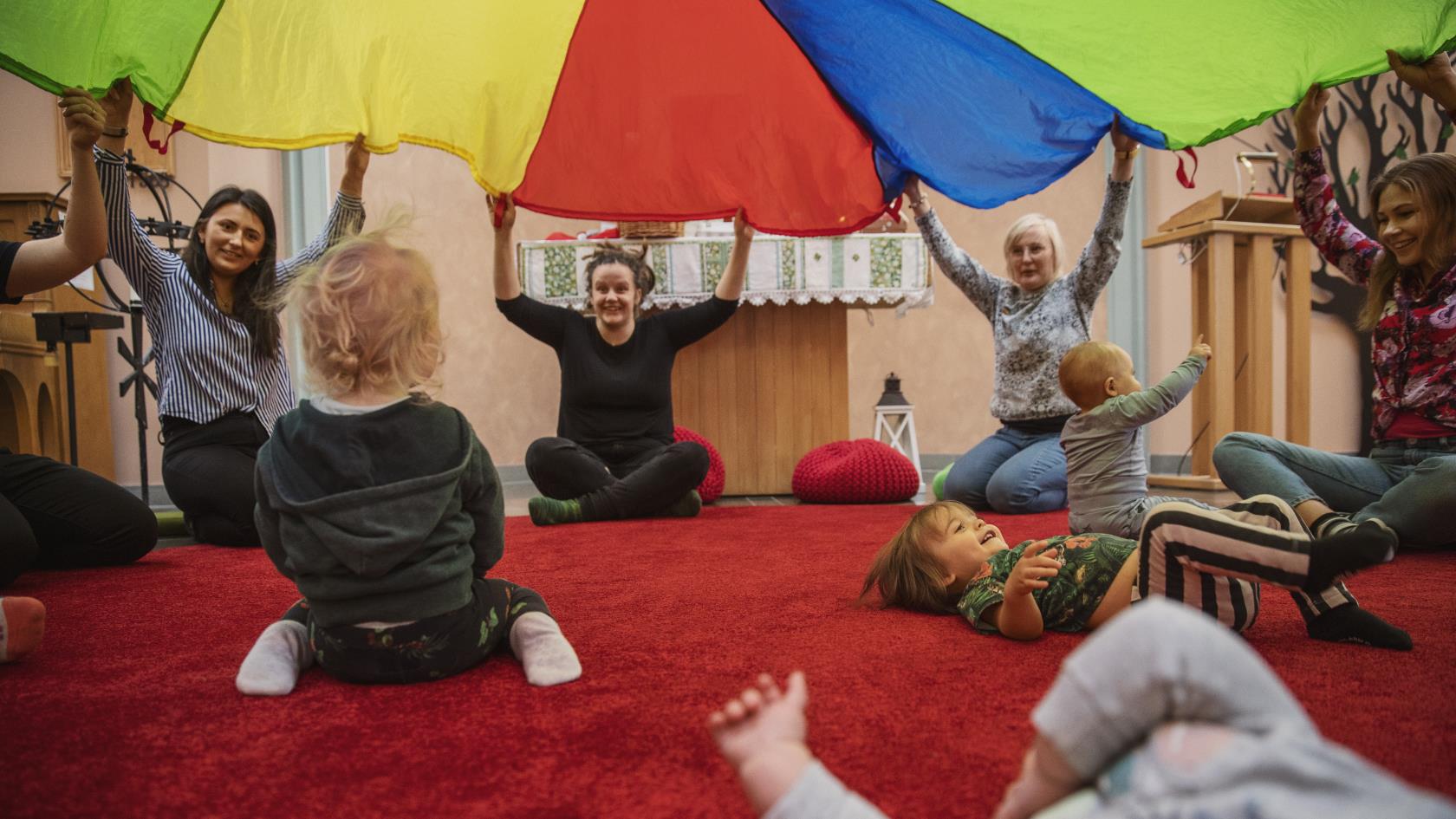 En grupp kvinnor sitter i en ring och lyfter tillsammans ett färgglatt tak. Inne i ringen, under taket, leker små bebisar.