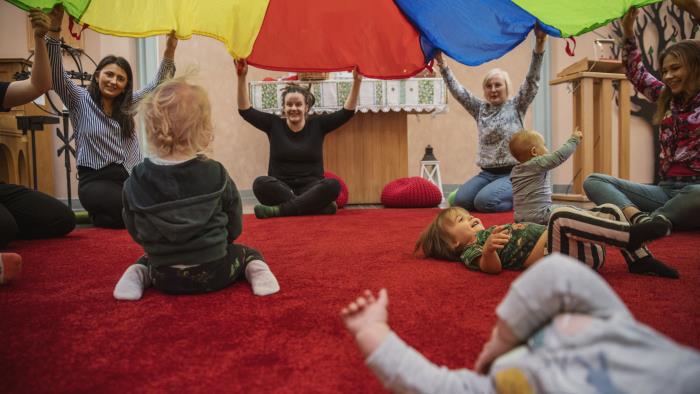 En grupp kvinnor sitter i en ring och lyfter tillsammans ett färgglatt tak. Inne i ringen, under taket, leker små bebisar.