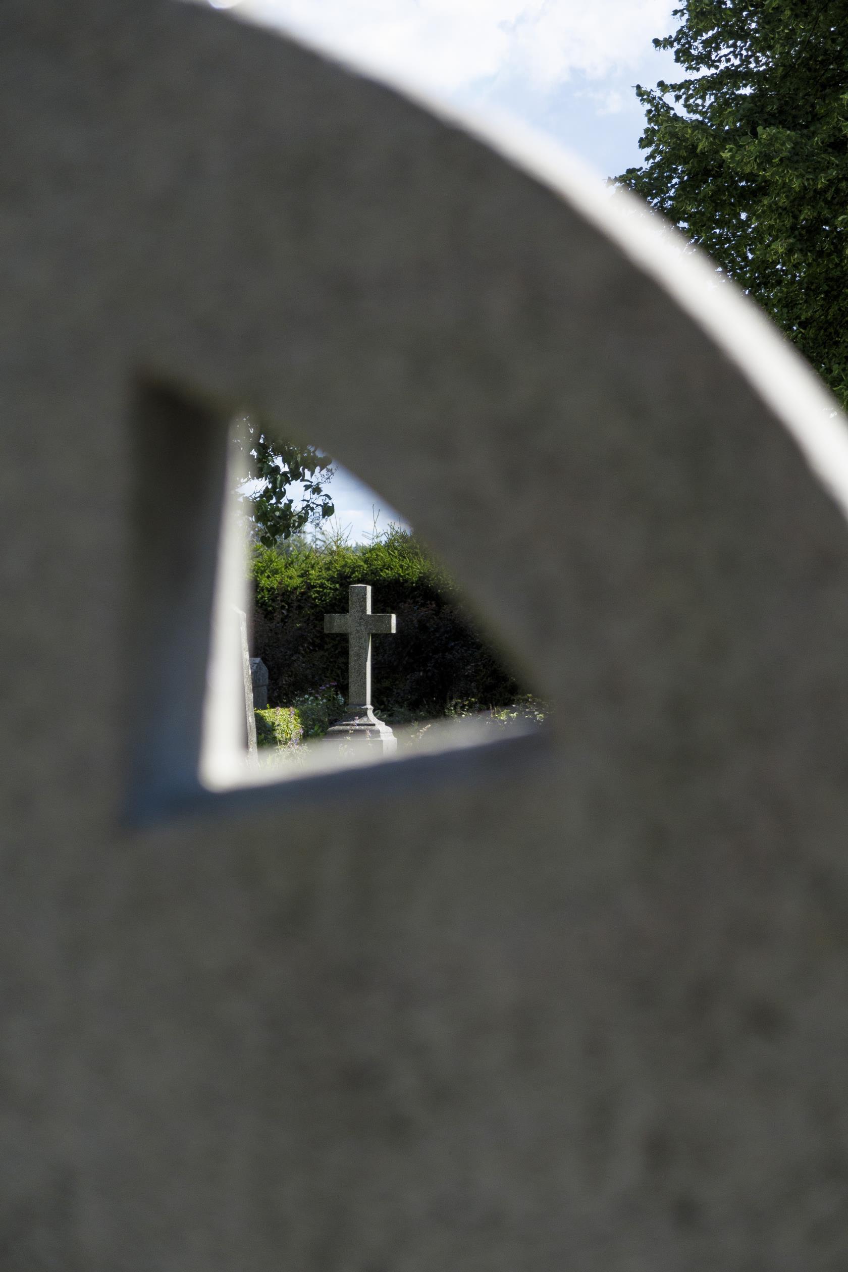 Genom ett hugget hål i en gravsten syns ett gravkors i sten längre bort.