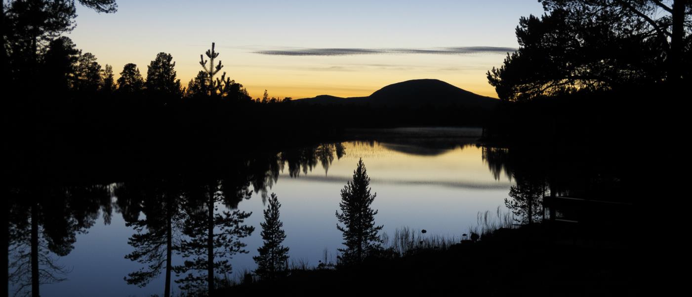 Skog och ett berg reflekteras på en stilla vattenyta i solnedgången.