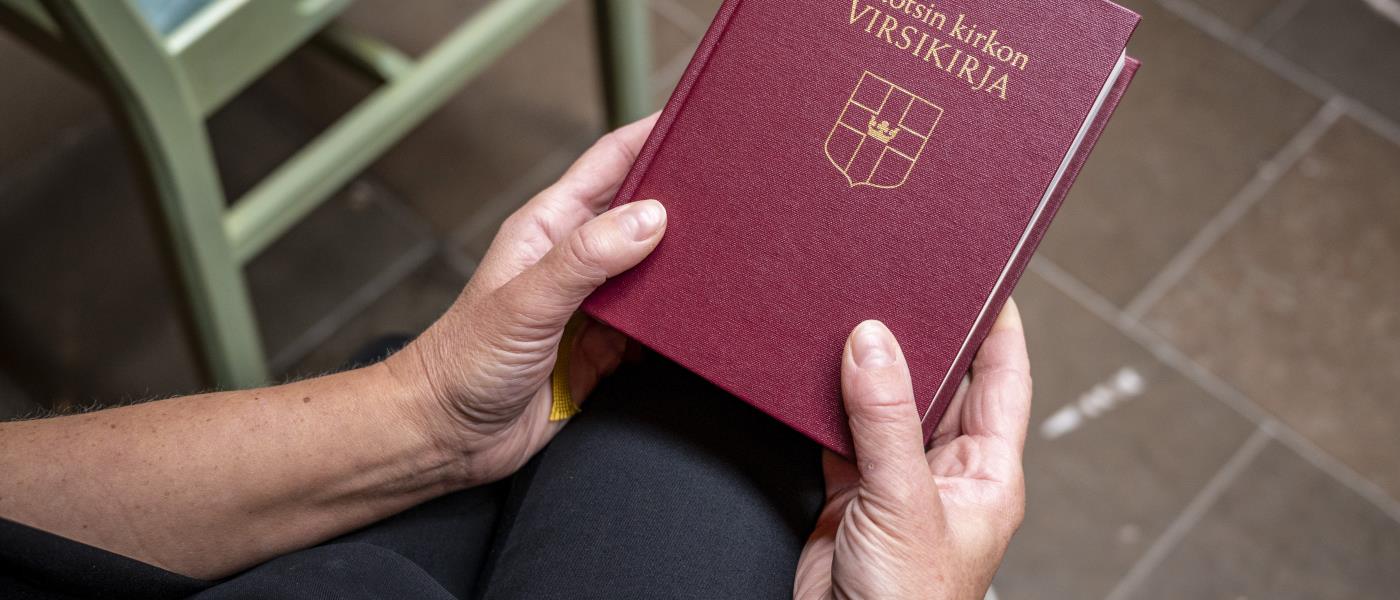 Någon sitter i ett kapell och håller en finsk psalmbok i händerna.