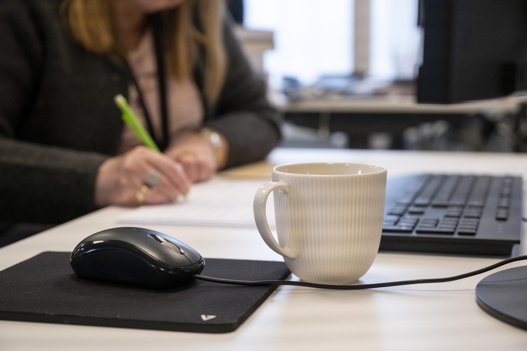 En kaffekopp står bredvid en datormus på ett skrivbord. En kvinna som antecknar syns suddigt i bakgrunden.