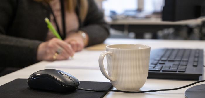 En kaffekopp står bredvid en datormus på ett skrivbord. En kvinna som antecknar syns suddigt i bakgrunden.