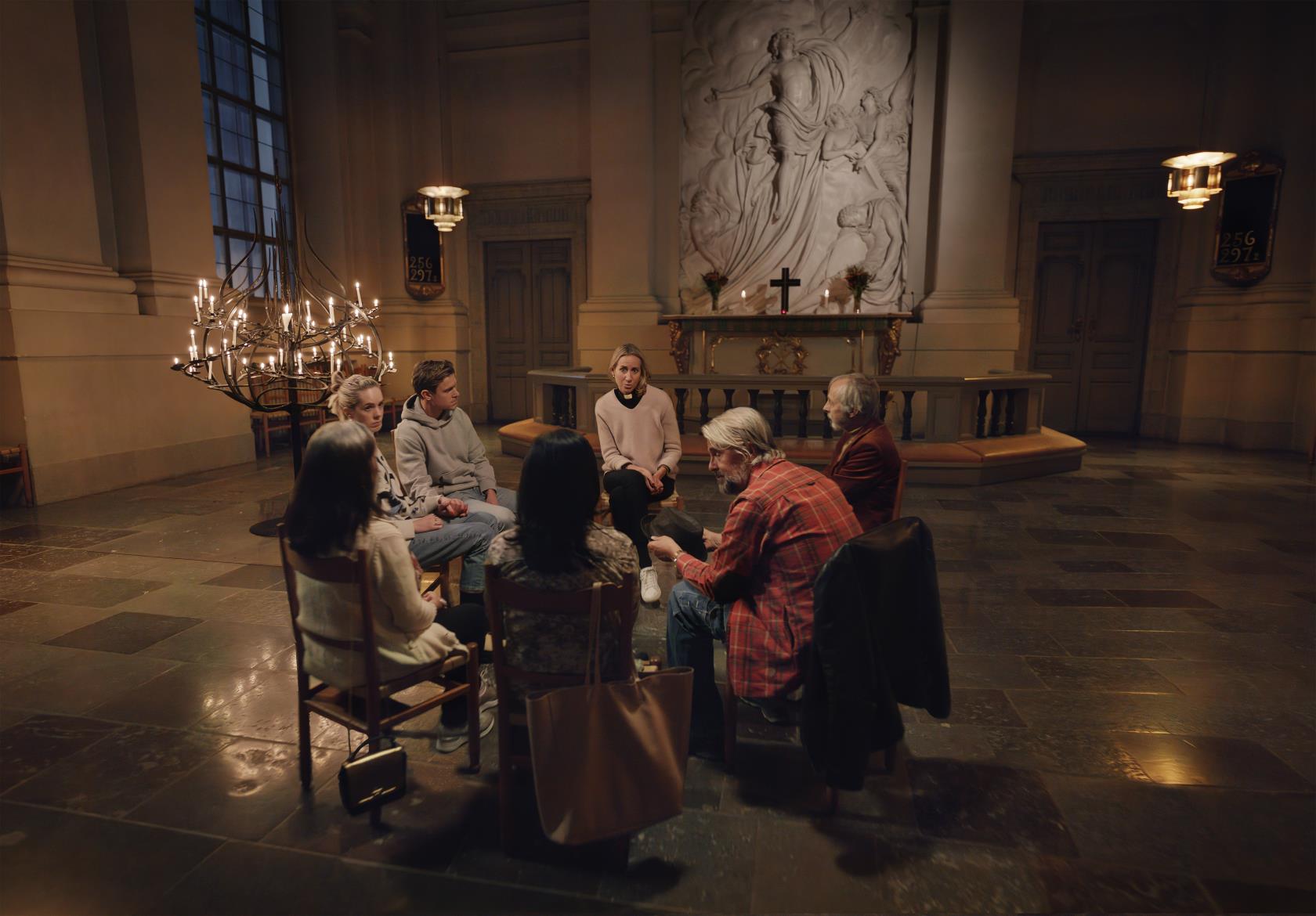 Några personer och en kvinnlig präst sitter på stolar i en ring i en kyrka.