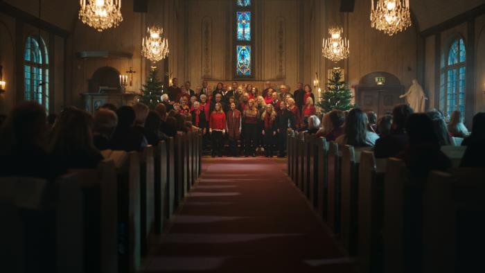 Mittgången i en fullsatt kyrka. Längst fram står en kyrkokör i julkläder mellan två julgranar och sjunger.