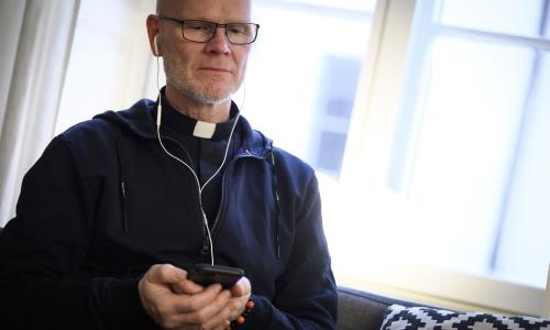 En manlig präst sitter i en soffa med en mobiltelefon i handen och hörlurar i öronen.