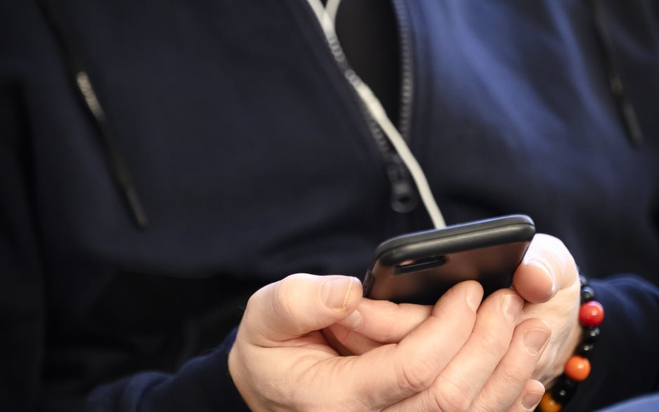 En manlig präst sitter i en soffa med en mobiltelefon i handen och hörlurar i öronen.