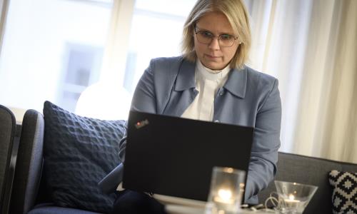 En kvinnlig präst i ljusblå kavaj sitter i en soffa med en laptop i knät och en mobiltelefon i handen.