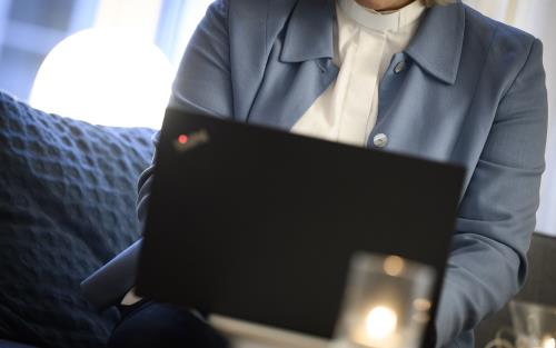 En kvinnlig präst i ljusblå kavaj sitter i en soffa med en laptop i knät och en mobiltelefon i handen.
