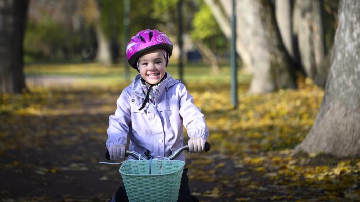 Ett barn med rosa hjälm cyklar genom en park full av höstlöv på marken.