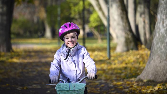 Ett barn med rosa hjälm cyklar genom en park full av höstlöv på marken.