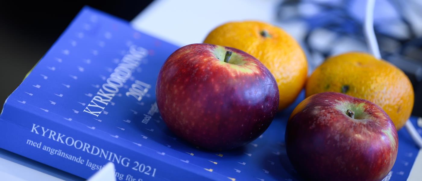 Två äpplen och två clementiner ligger på en blå bok med titeln Kyrkoordning 2021.