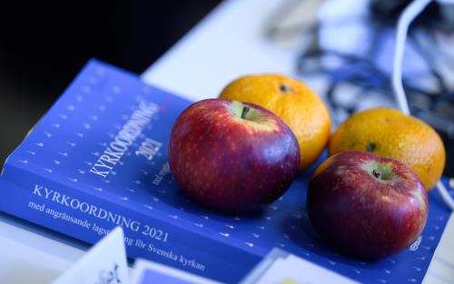 Två äpplen och två clementiner ligger på en blå bok med titeln Kyrkoordning 2021.