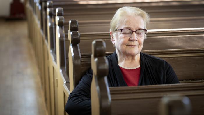 En äldre dam sitter med slutna ögon i de tomma kyrkbänkarna.